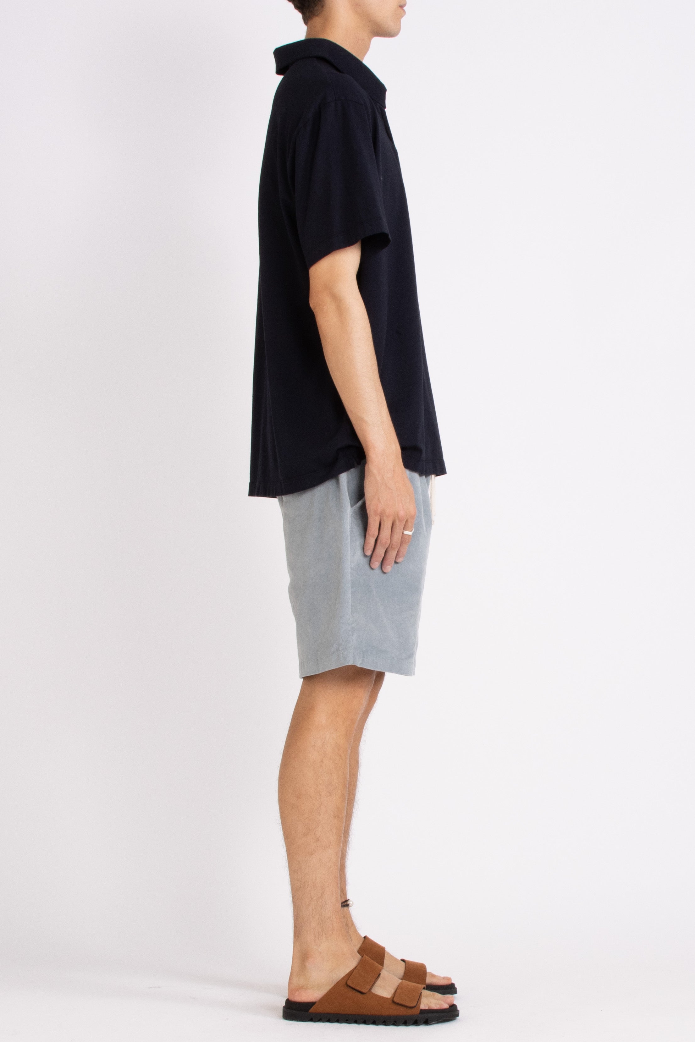 model wearing navy blue cotton polo shirt, light blue shorts and tan slides on a white background, full side view