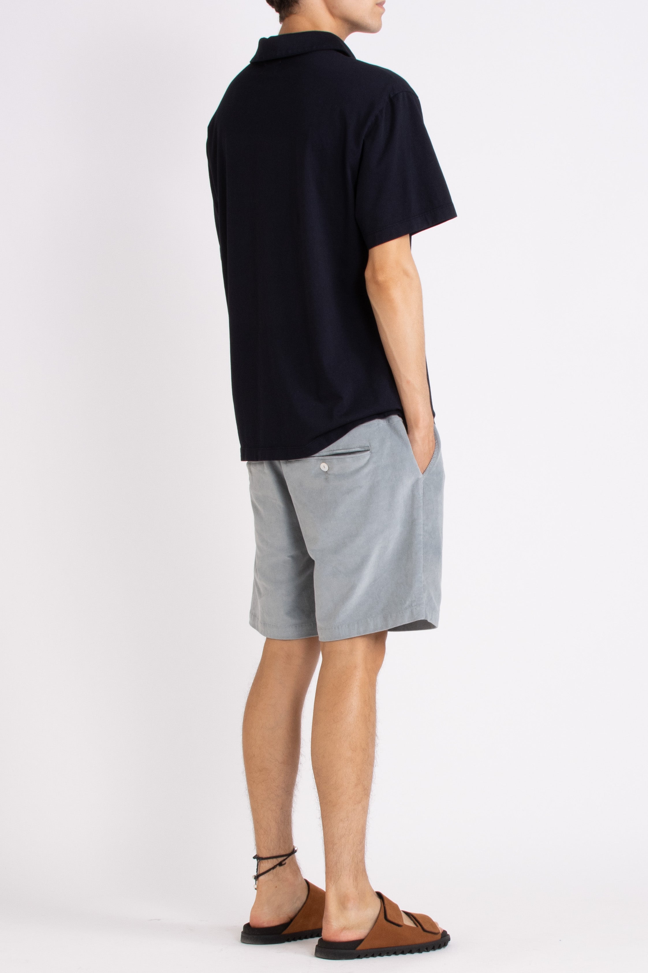 model wearing navy blue cotton polo shirt, light blue shorts and tan slides on a white background, side back view