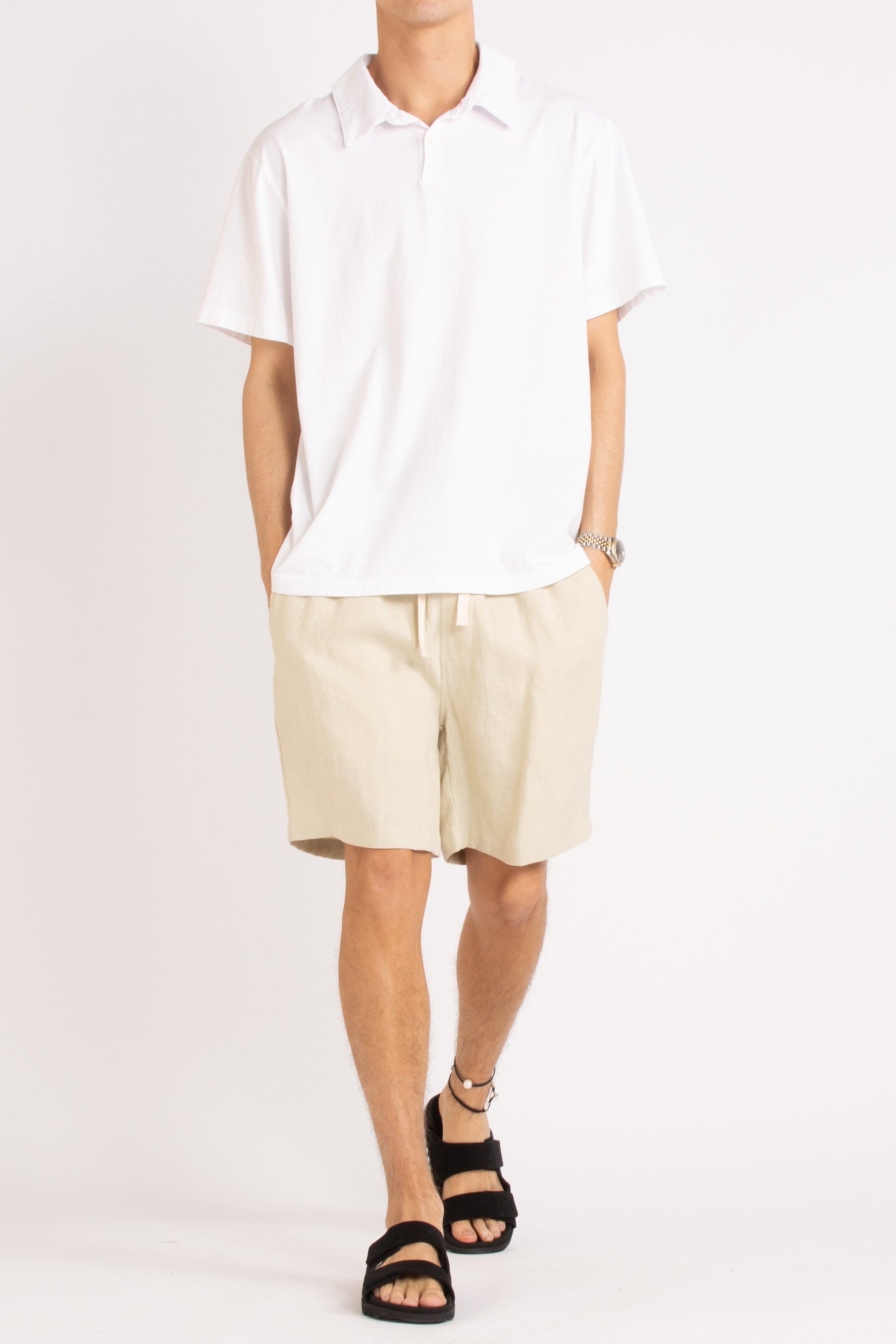 model wearing white cotton polo shirt, natural linen shorts and black slides on a white background, front view