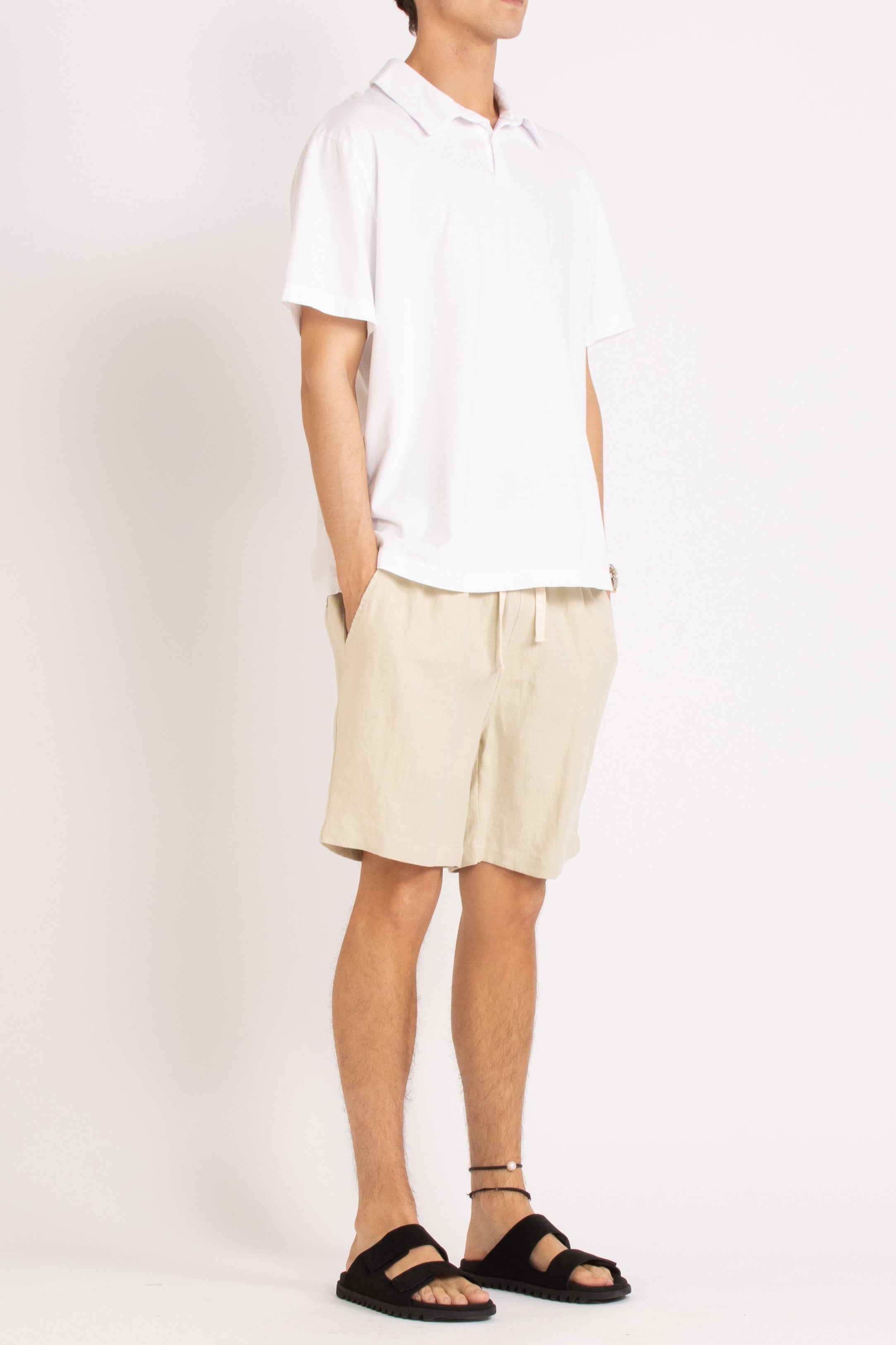 model wearing white cotton polo shirt, natural linen shorts and black slides on a white background, front side view