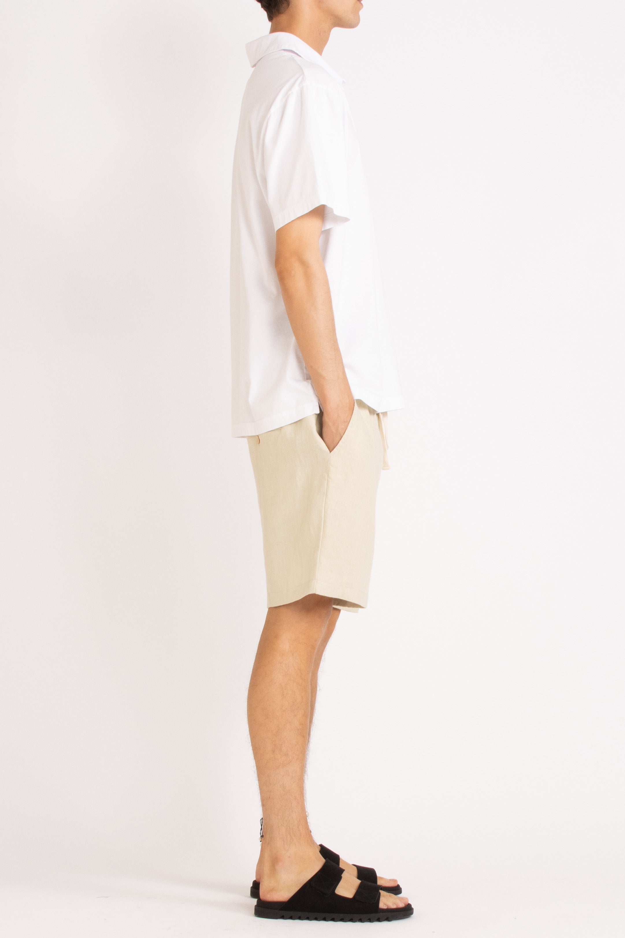 model wearing white cotton polo shirt, natural linen shorts and black slides on a white background, side view