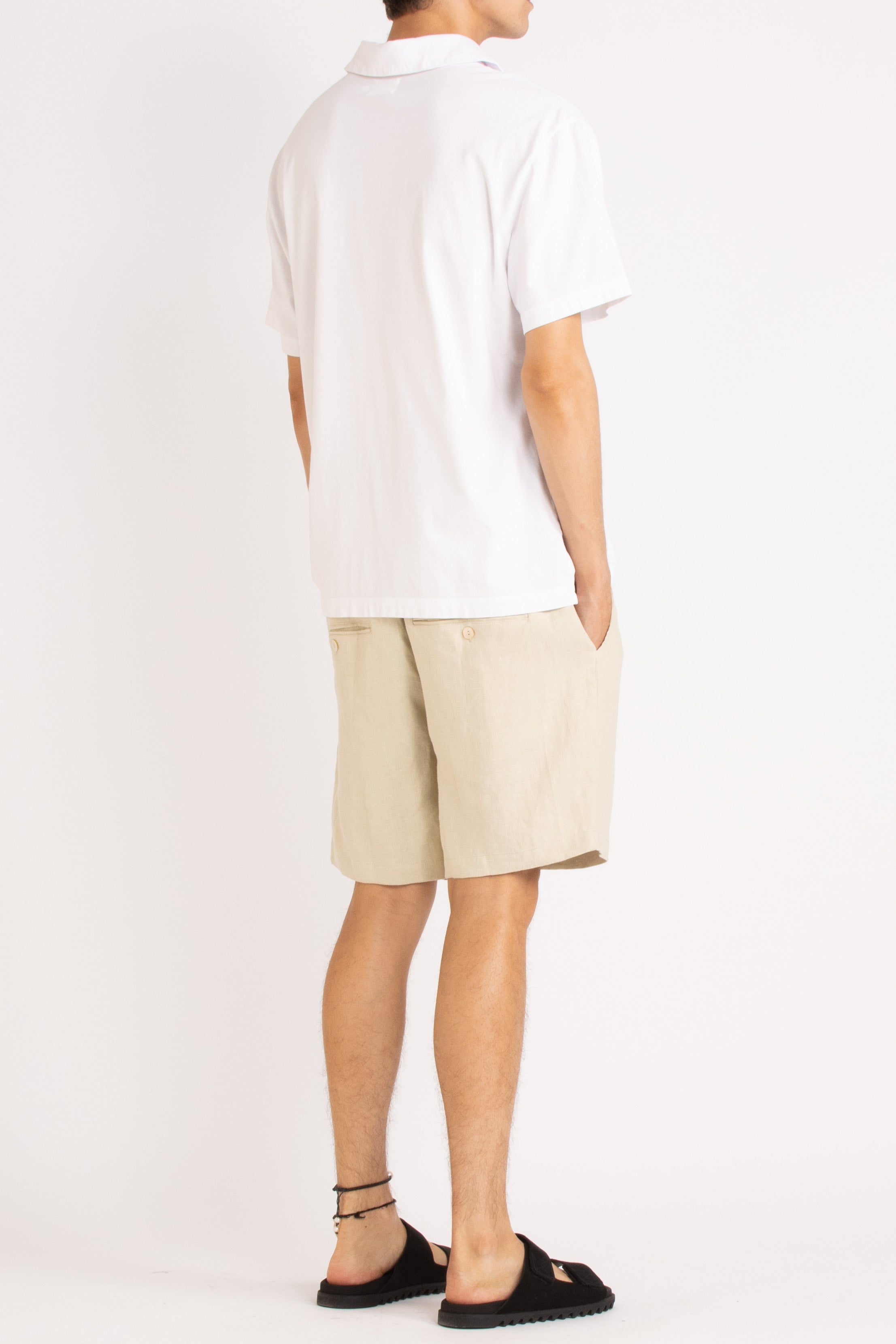 model wearing white cotton polo shirt, natural linen shorts and black slides on a white background, back side view
