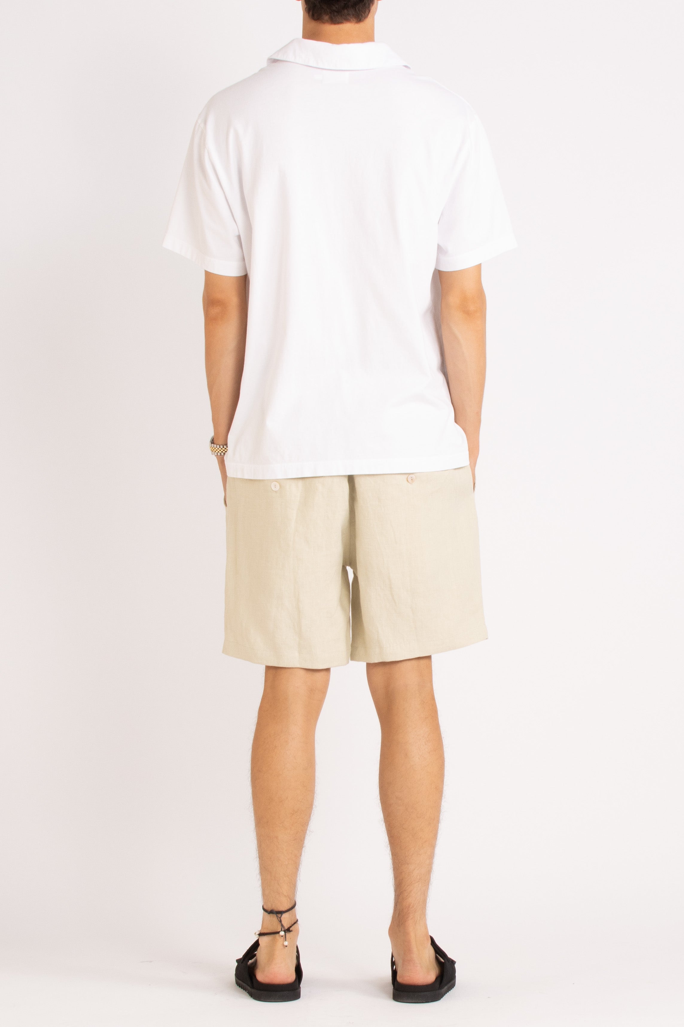 model wearing white cotton polo shirt, natural linen shorts and black slides on a white background, back view