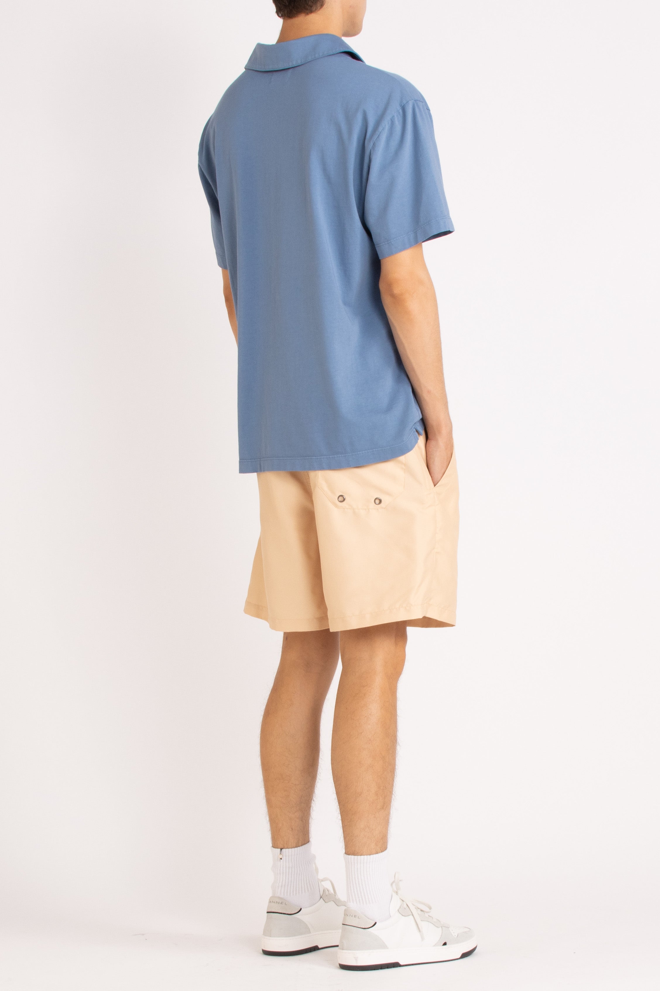 model wearing blue cotton polo shirt, light sand shorts and white sneakers on a white background, back side view