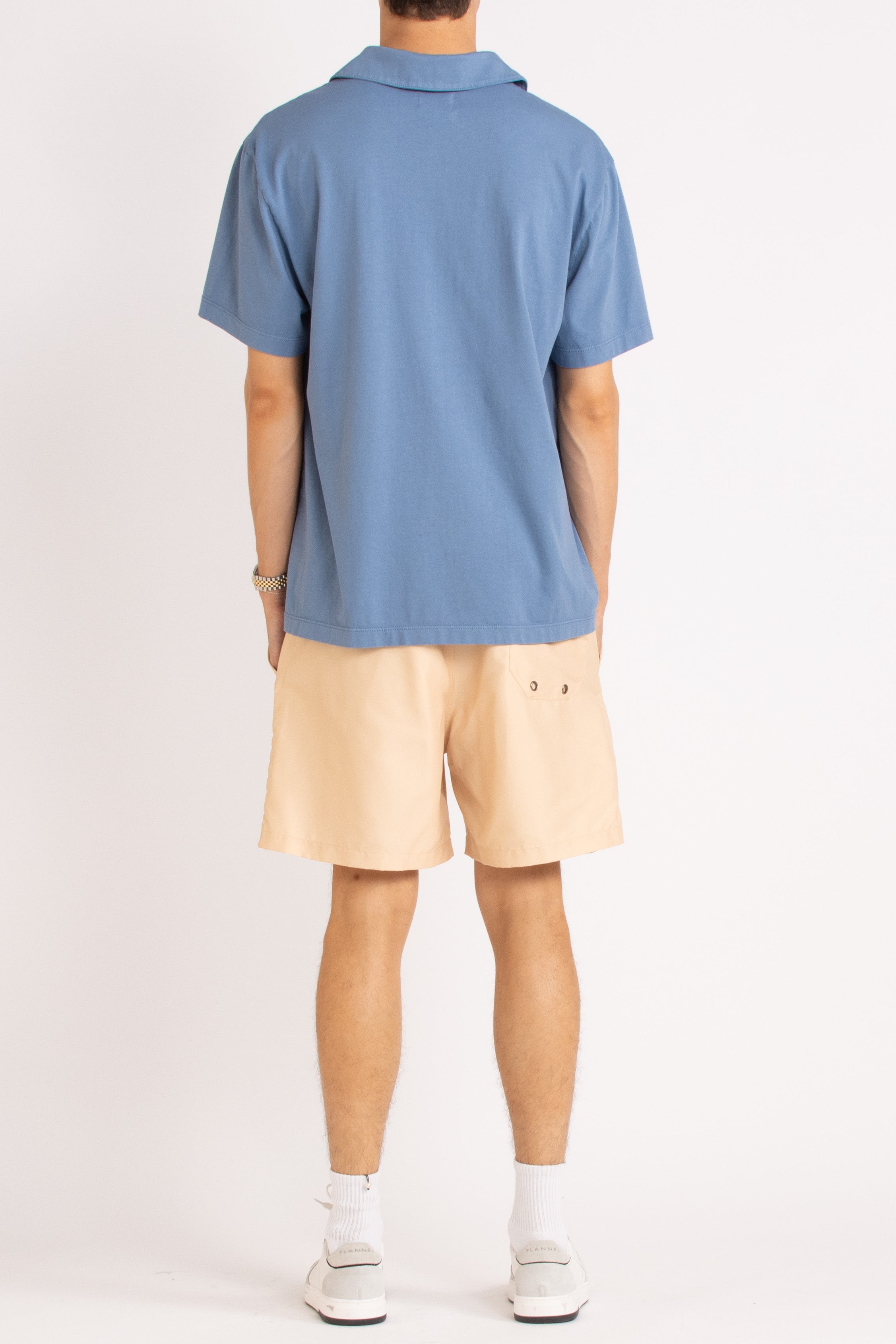 model wearing blue cotton polo shirt, light sand shorts and white sneakers on a white background, back view