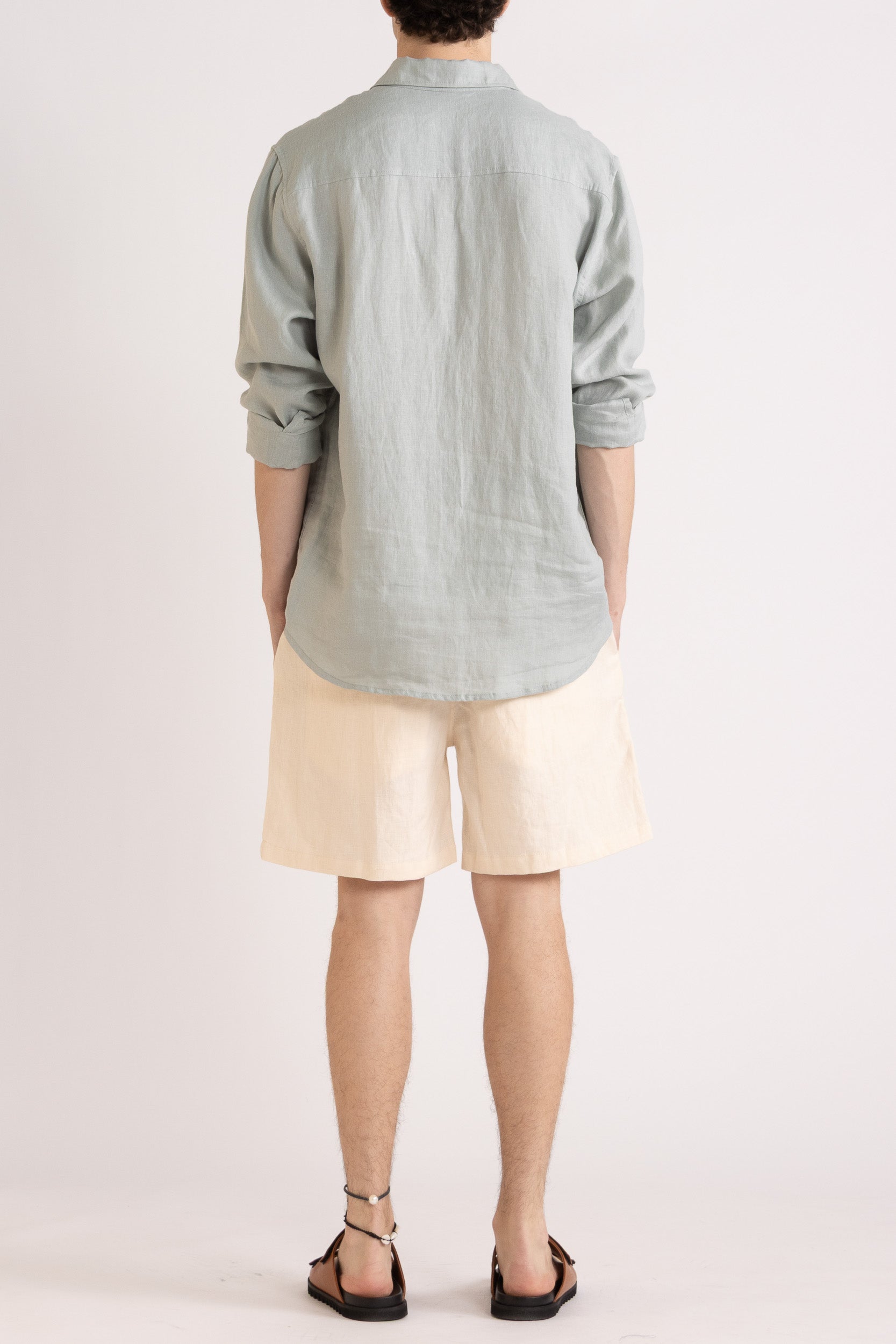 Person wearing a light blue long sleeve linen button-up shirt and natural linen shorts on a white background, back view.