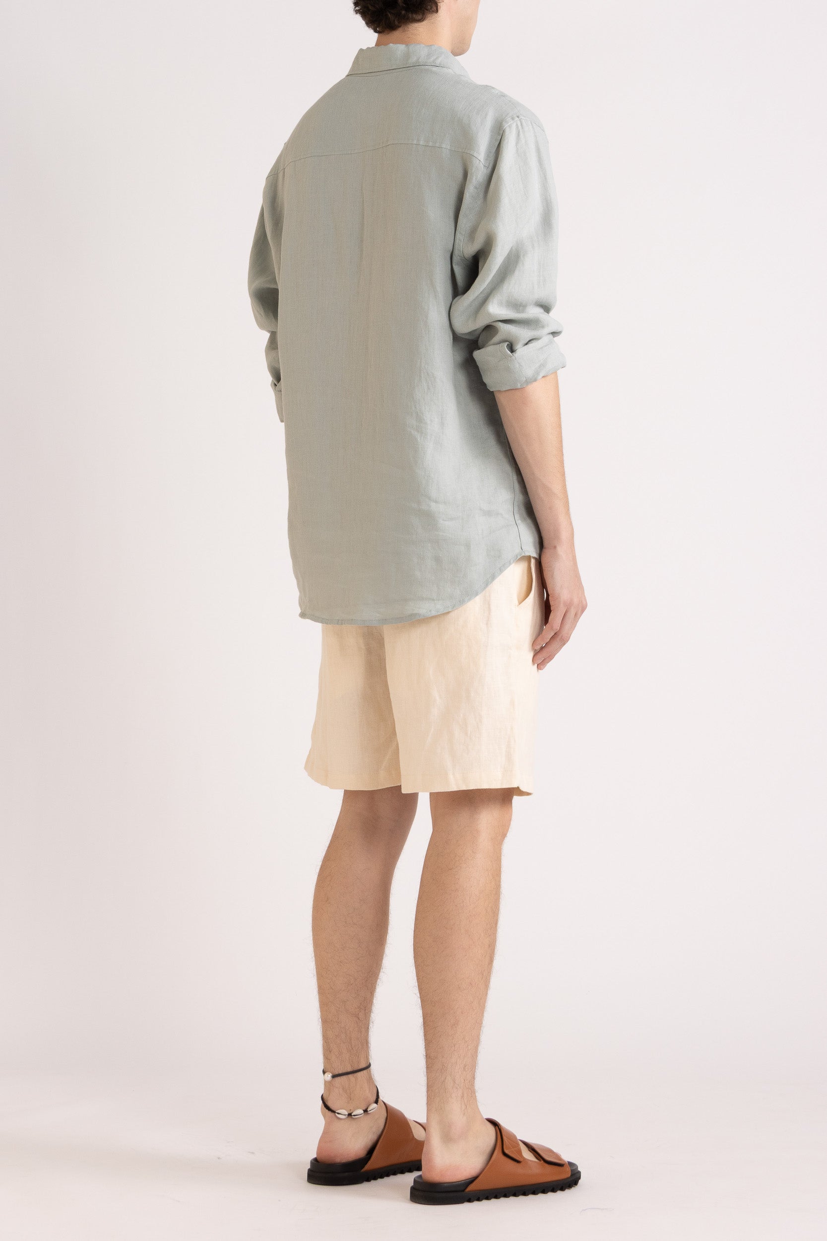 Person wearing a light blue long sleeve linen button-up shirt and natural linen shorts on a white background, side/back view