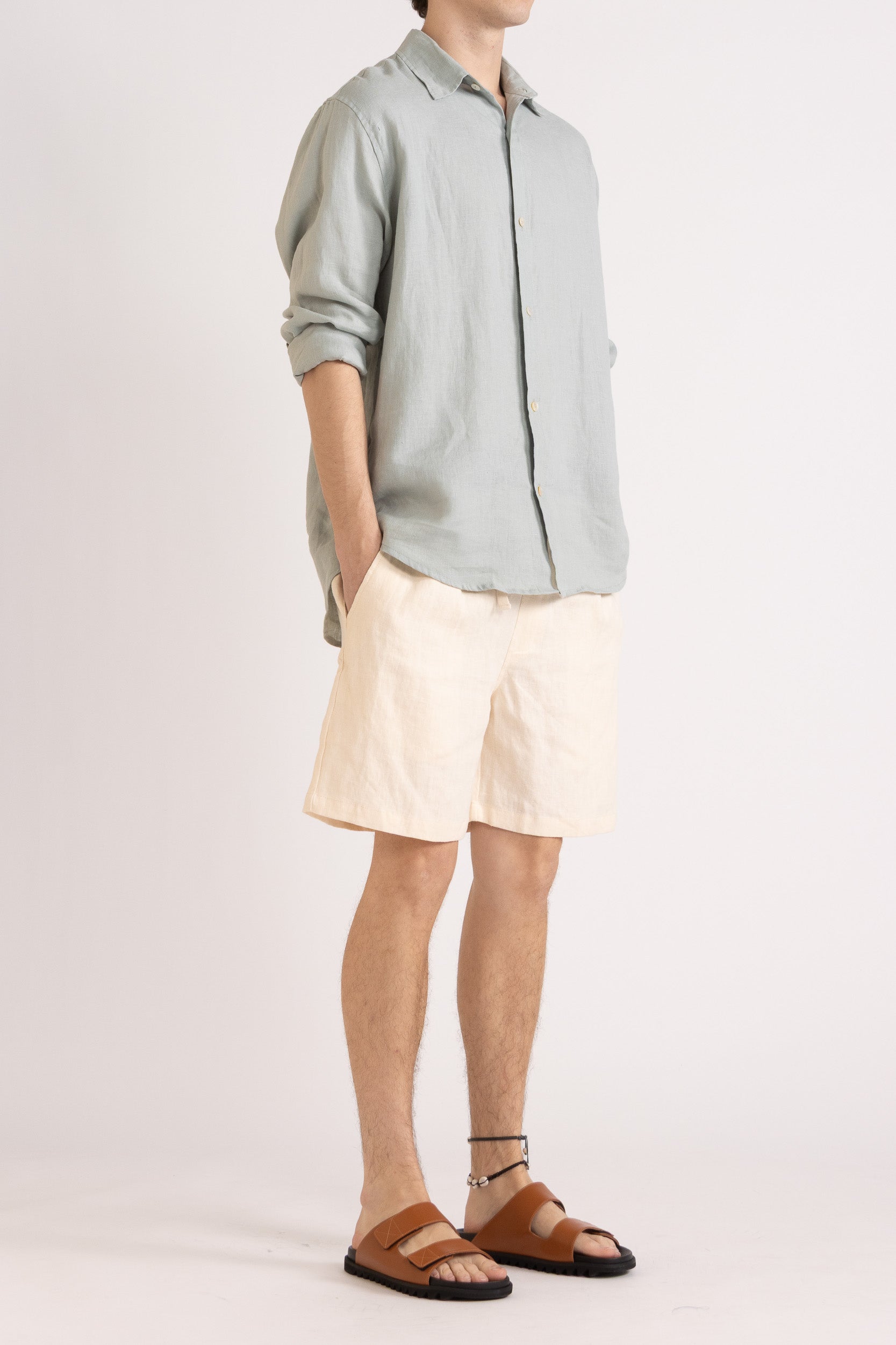 Person wearing a light blue long sleeve linen button-up shirt and natural linen shorts on a white background, front/side view