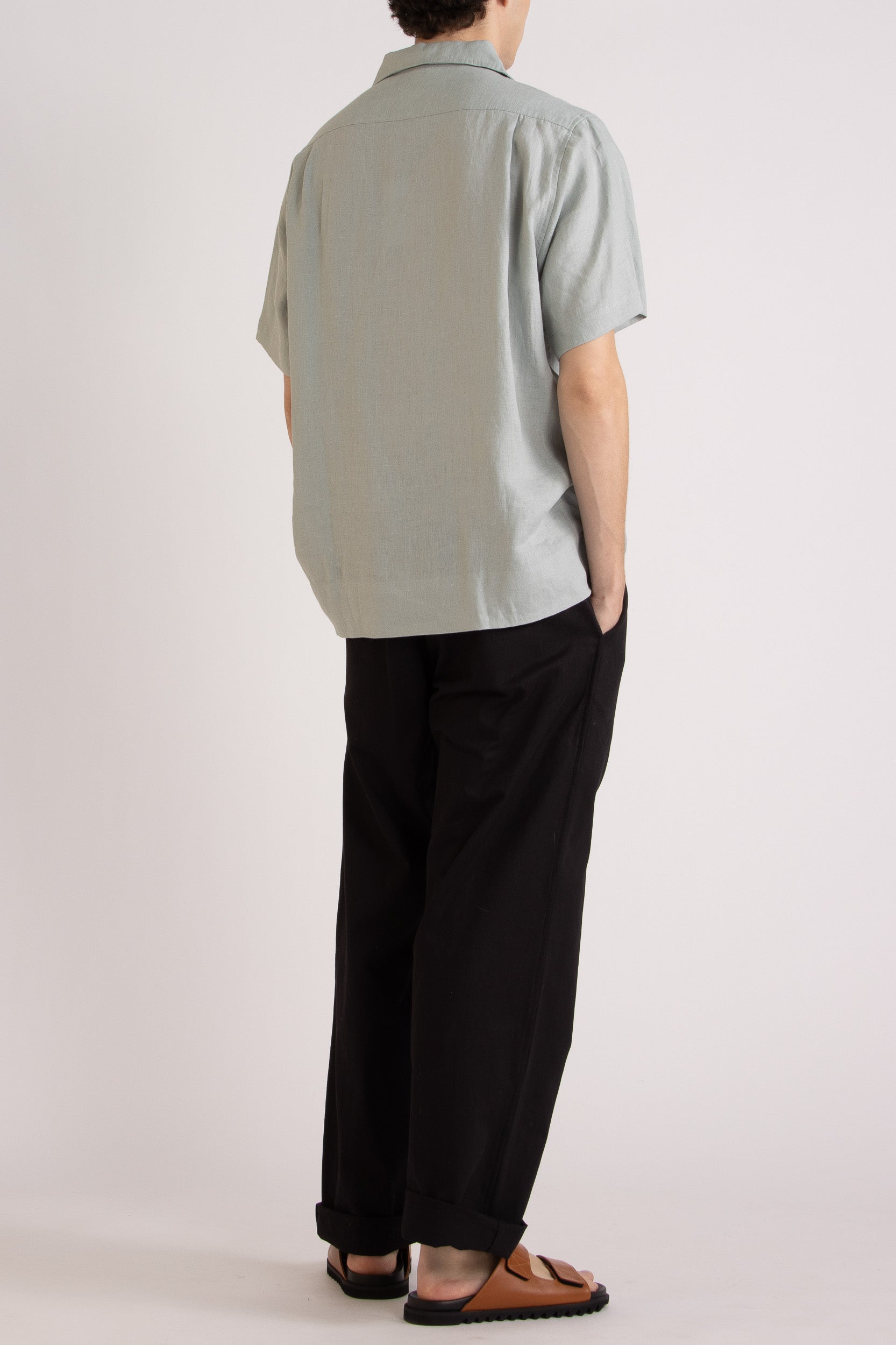 Model wearing a light blue linen short sleeve shirt and black linen pants on a white background, back/side view