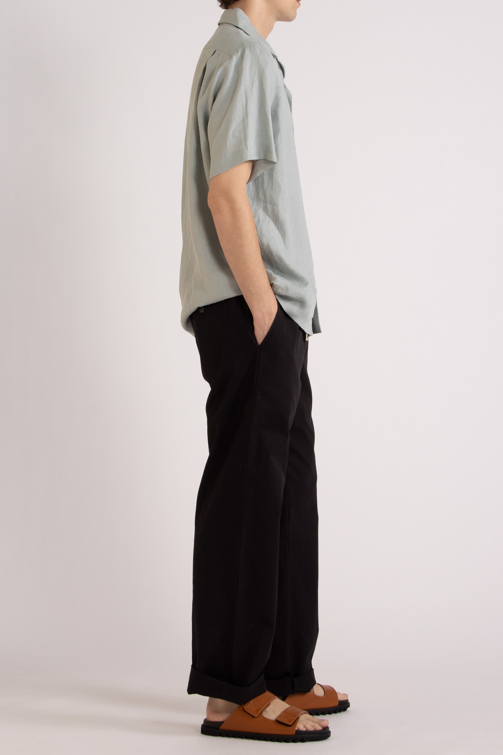 Model wearing a light blue linen short sleeve shirt and black linen pants on a white background, side view