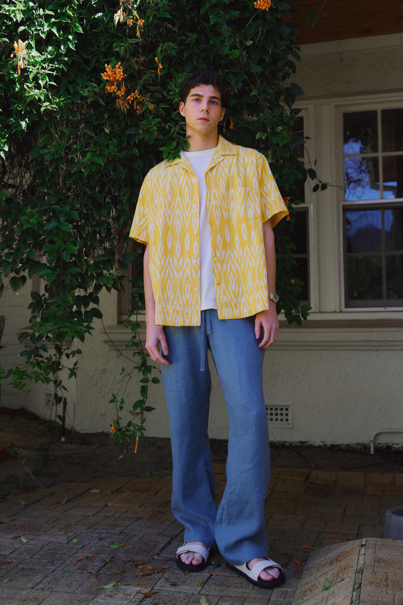 Model wearing a yellow cotton patterned shirt open with white t-shirt under and blue linen pants, campaign image