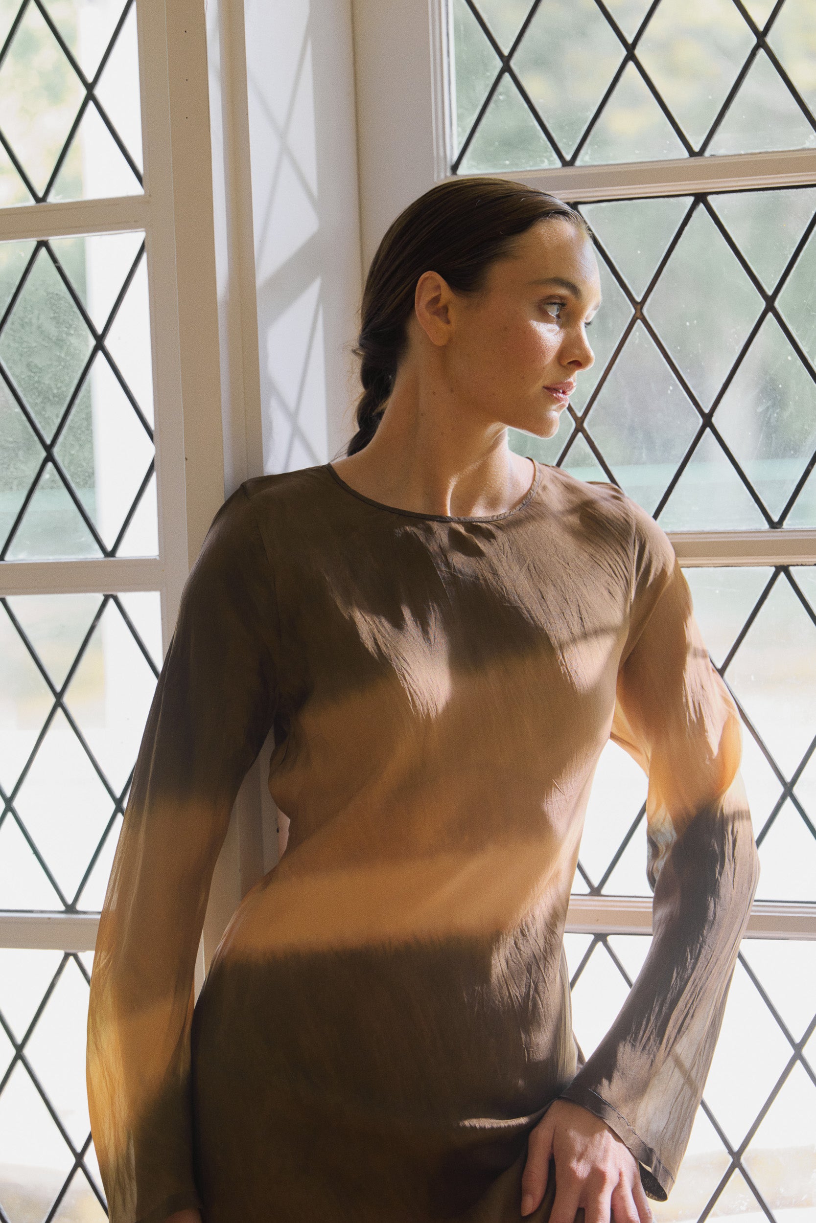 Woman wearing a silk habotai long sleeve dress dip dyed in earthy tones, looking out of a window