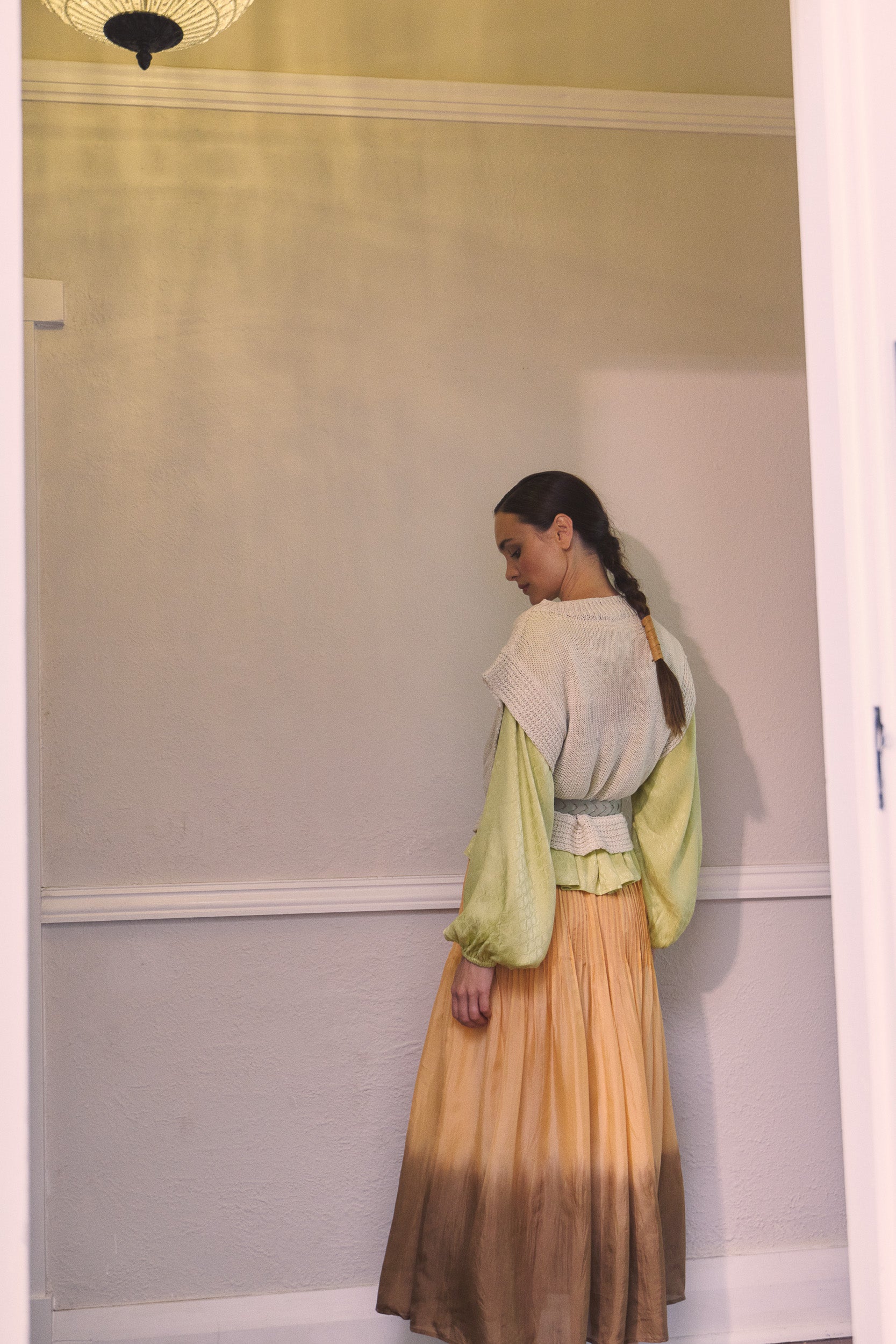 Woman wearing oat linen vest layered over a vanilla long sleeve top, paired over a light orange dip dyed maxi skirt with a cream braided belt.