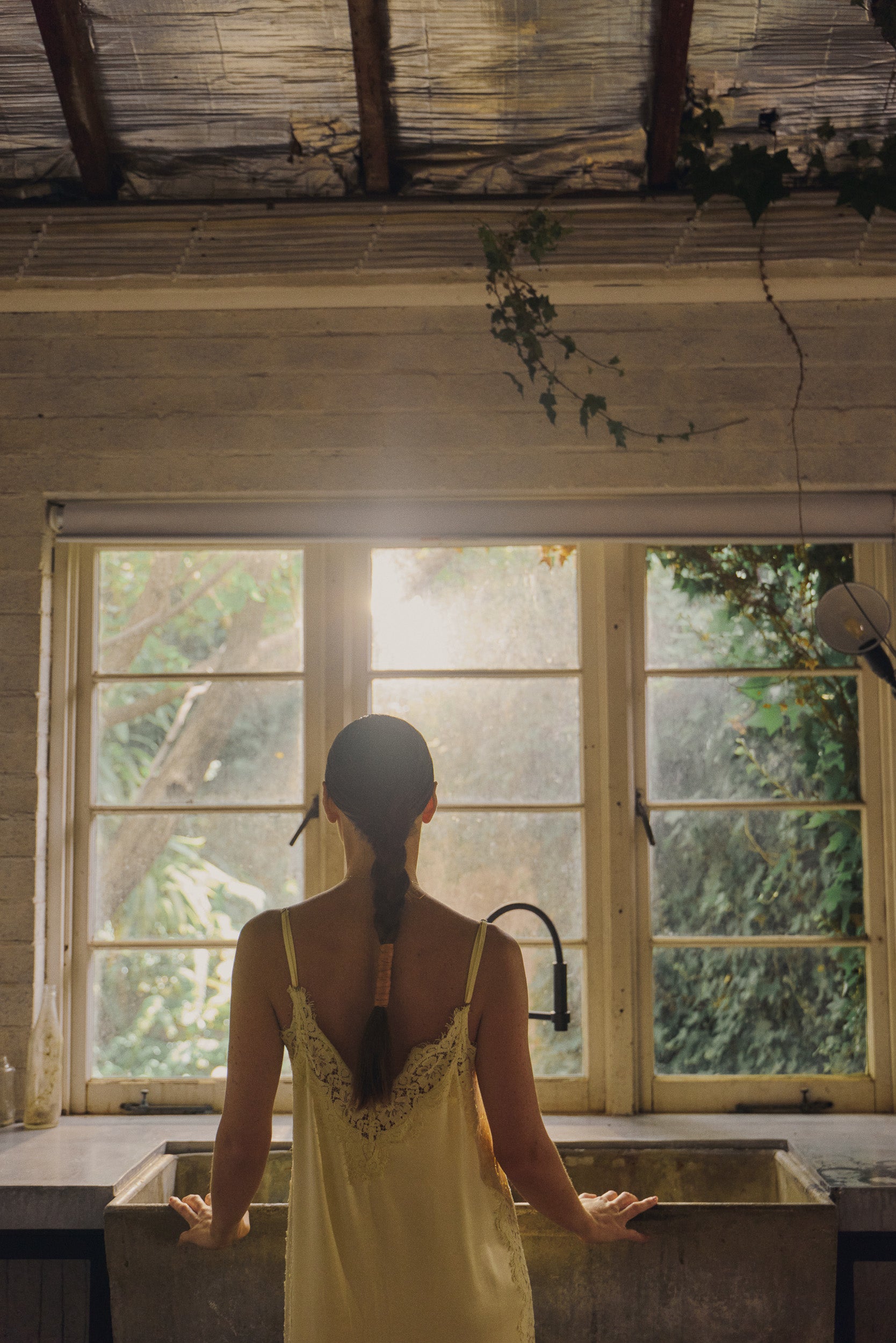 Woman wearing vanilla lace-trimmed camisole slip dress looking out of a window.