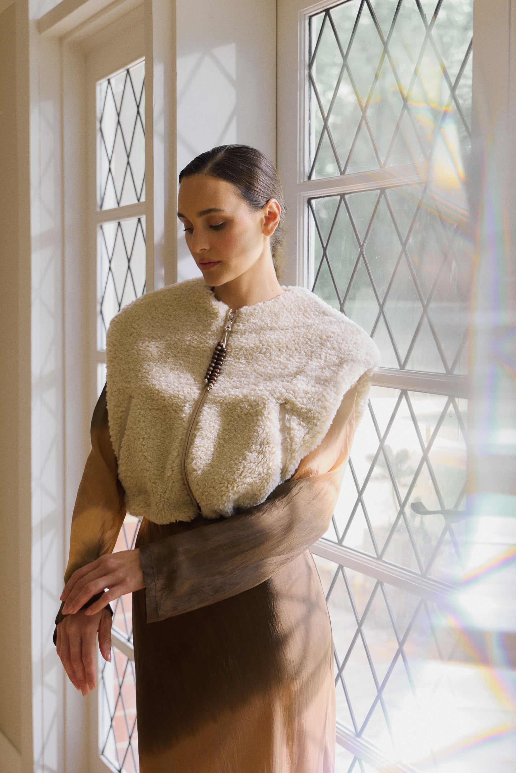 Woman wearing oat coloured faux fur vest over a long sleeve silk habotai maxi dress dip dyed in earthy tones, standing next to a window