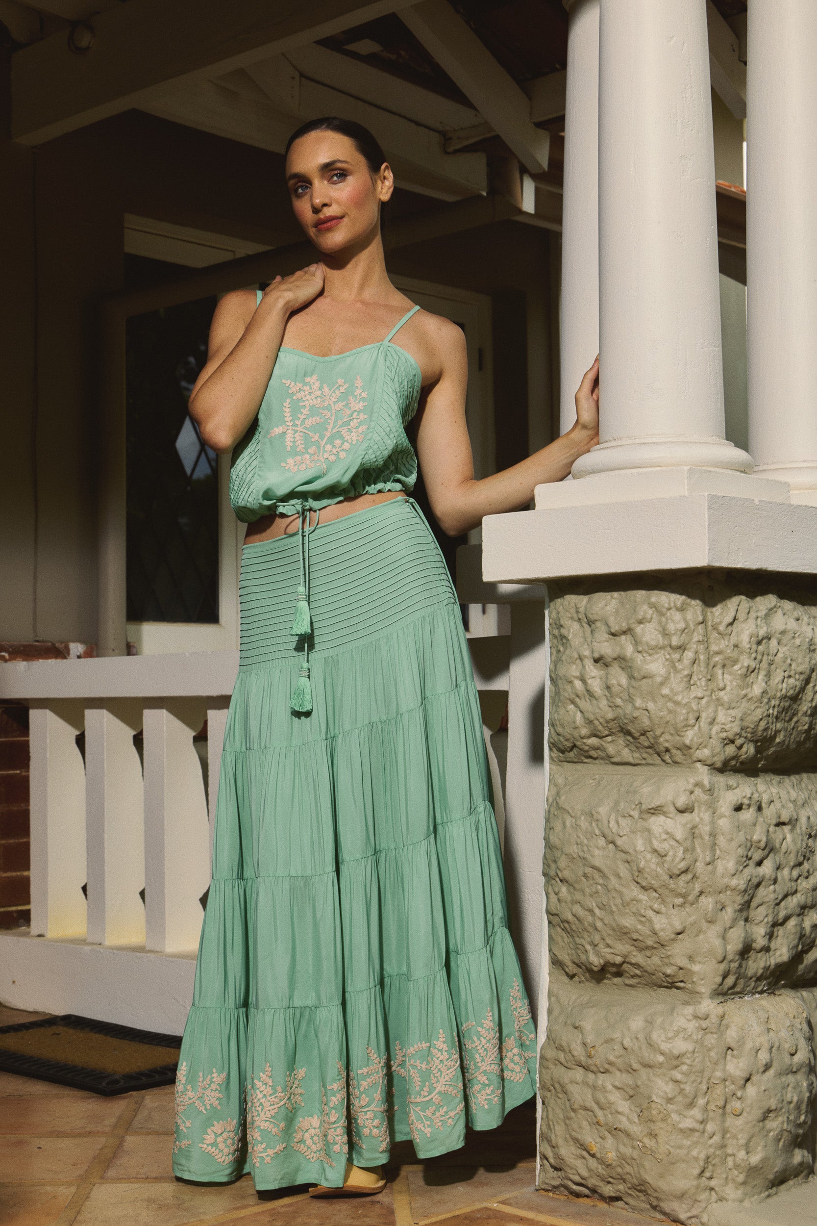 Woman in a mint green camisole and matching long tiered skirt with white floral embroidered standing on a stone porch.