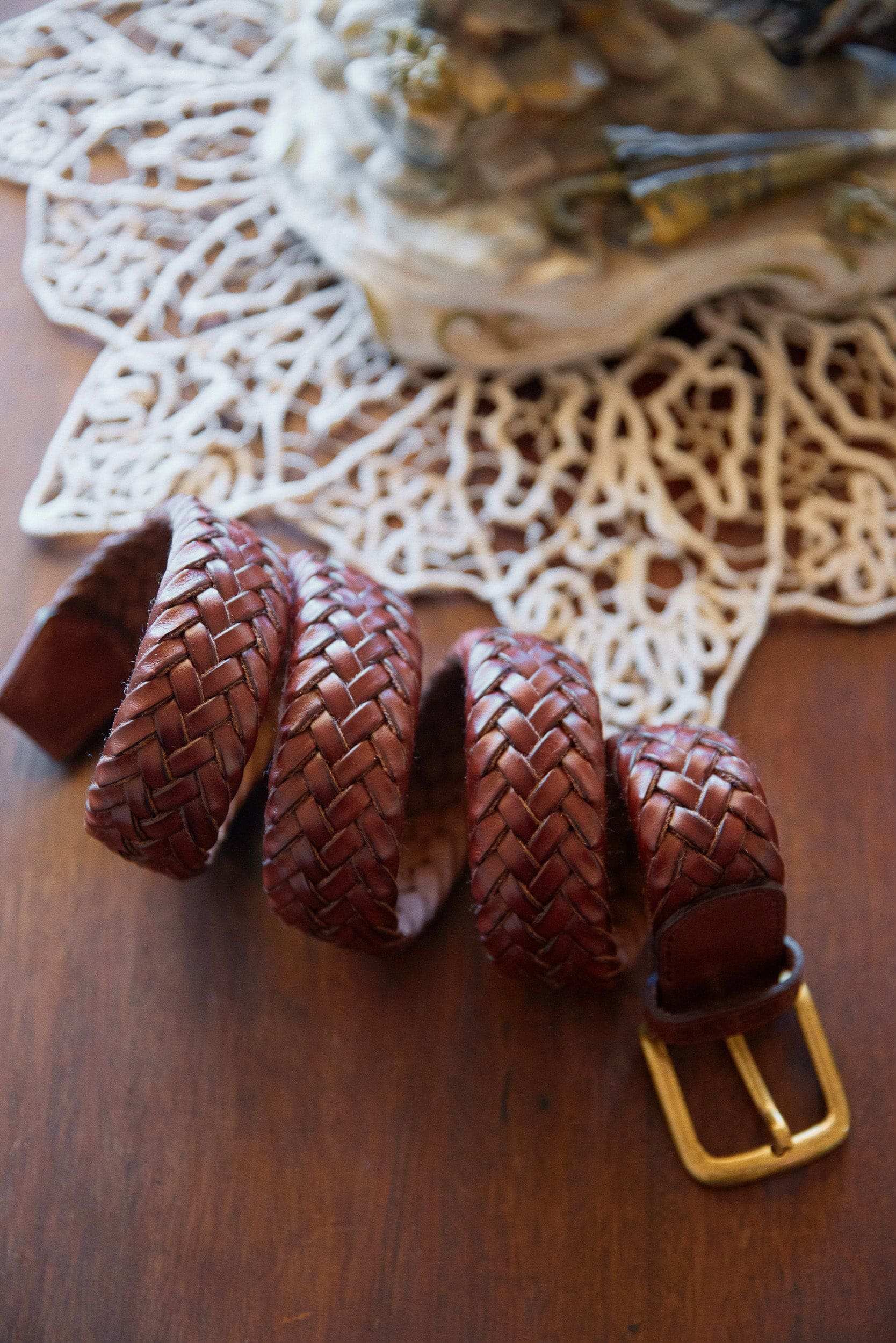 Brown woven leather belt with a gold buckle on a wooden surface with a doily in the background.