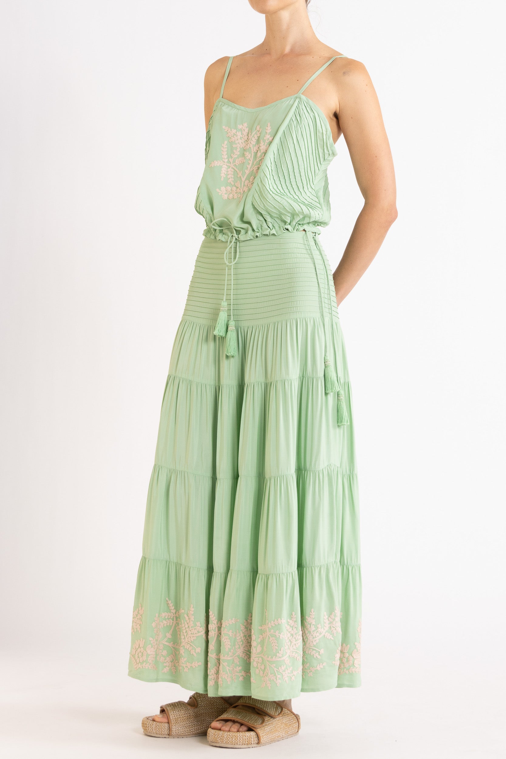 Woman wearing a mint green camisole and long, tiered skirt with white floral embroidery on a white background