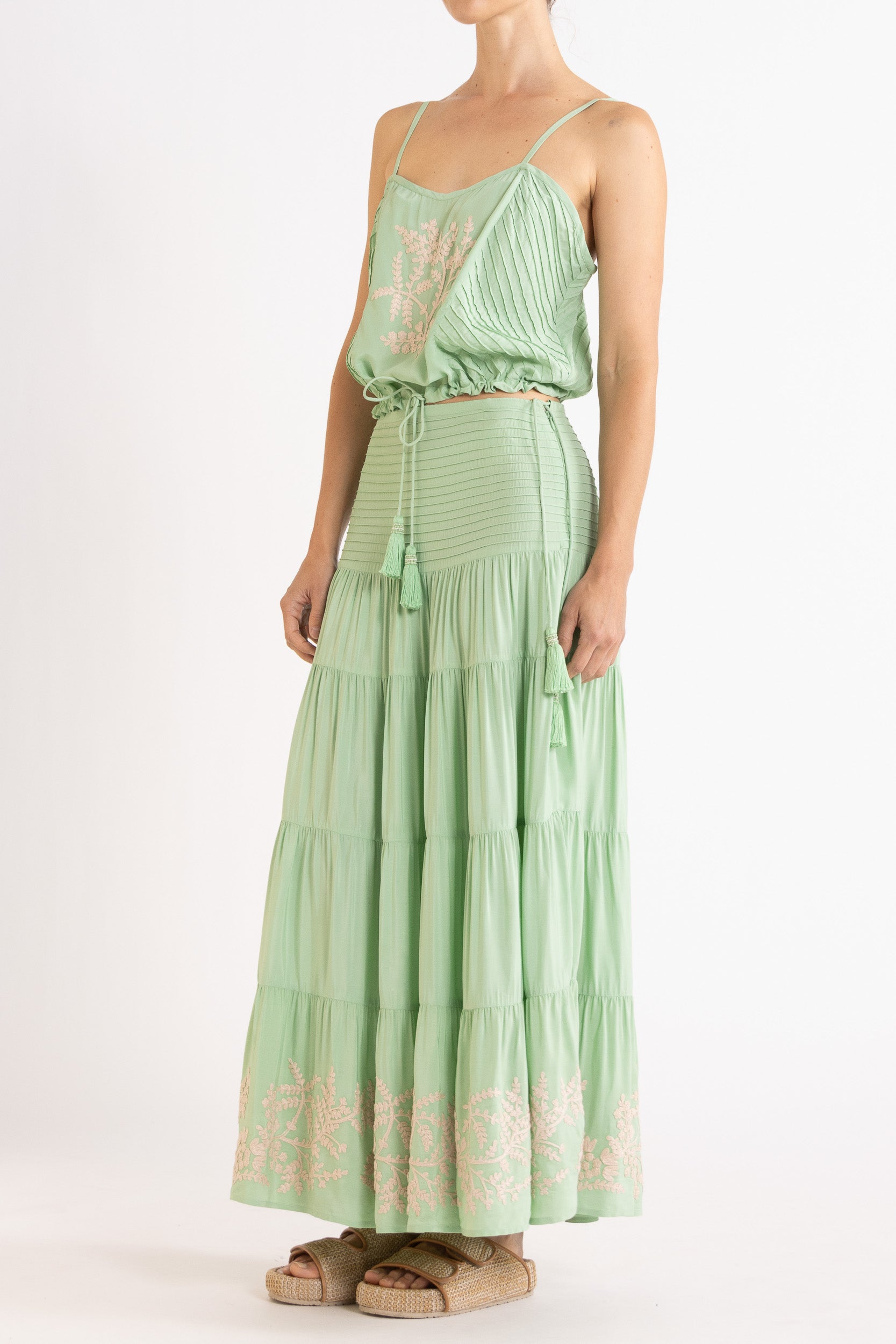 Woman wearing a mint green camisole and long, tiered skirt with white floral embroidery on a white background