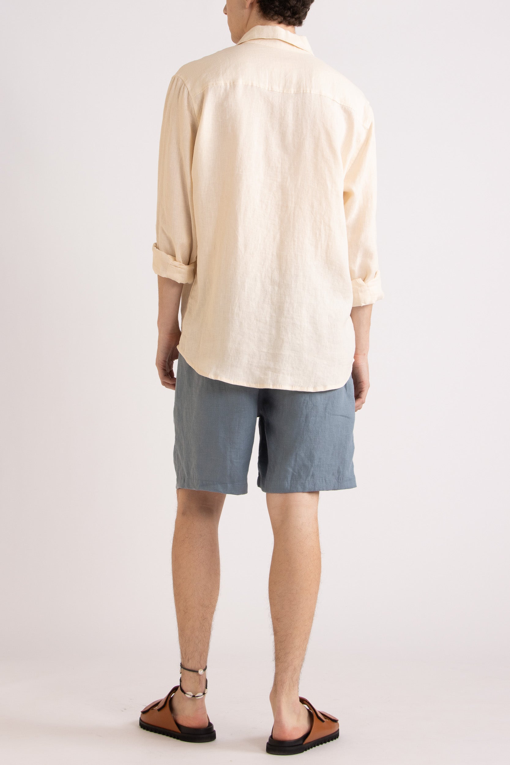 Model wearing a natural linen long sleeve shirt and steel blue linen shorts on a white background, back view.