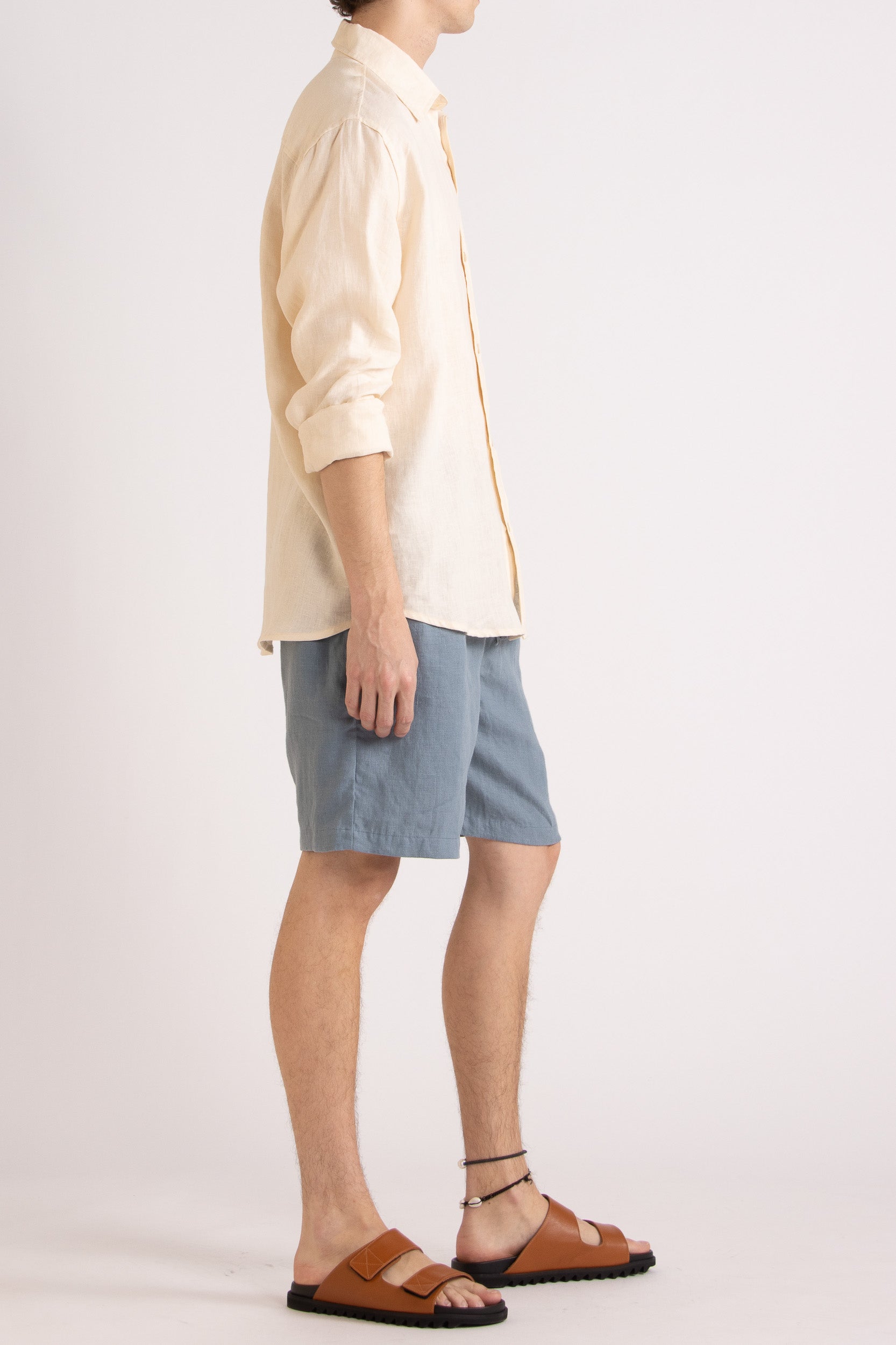 Model wearing a natural linen long sleeve shirt and steel blue linen shorts on a white background, side view.