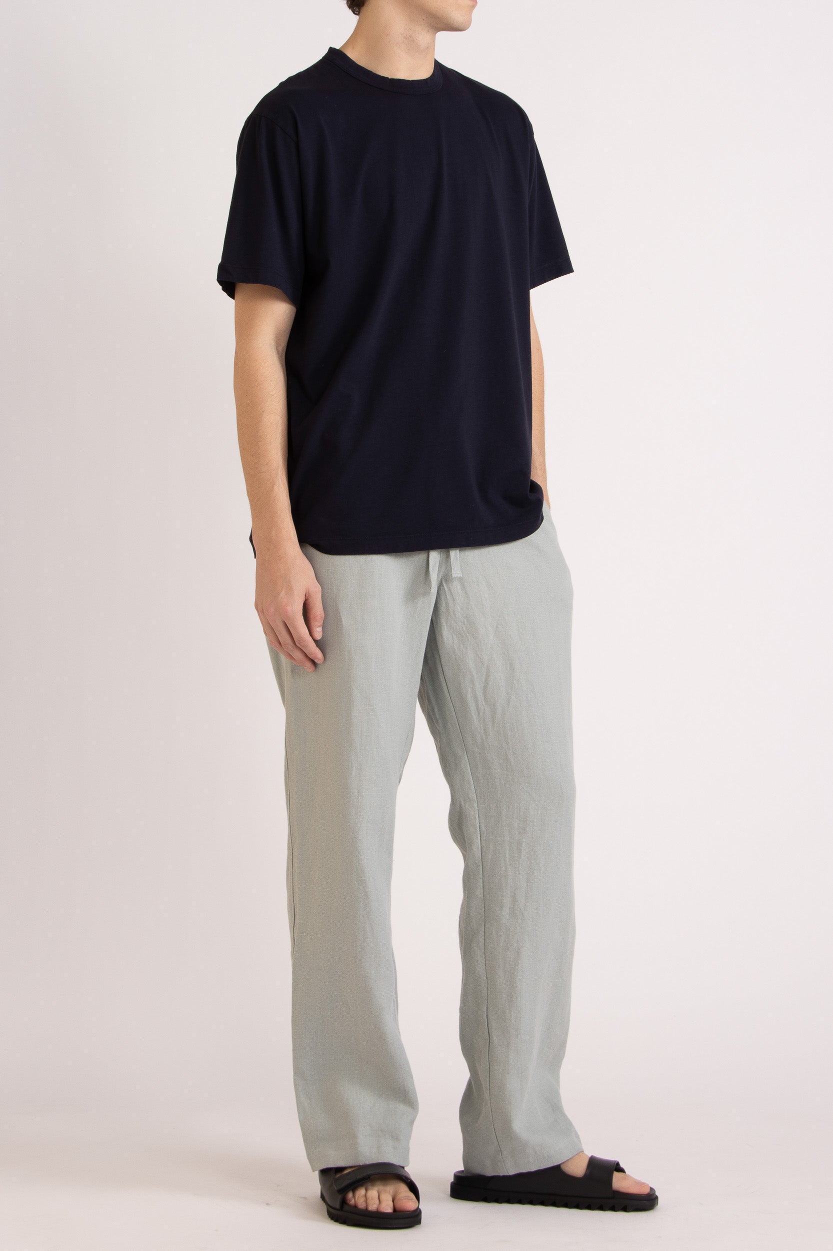 Man wearing a dark navy t-shirt, mint linen pants and black leather slides on a white background
