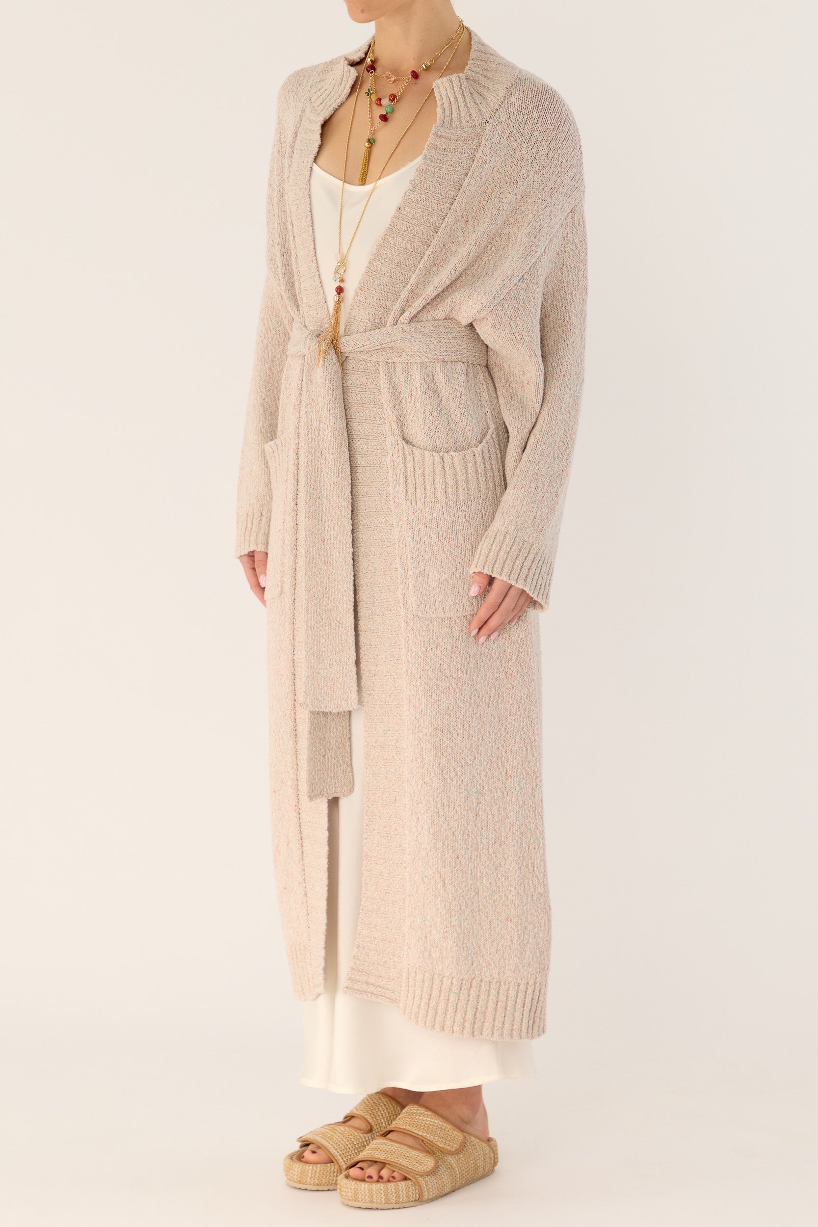 Model wearing beige knitted longline cardigan and white silk slip, front-side view