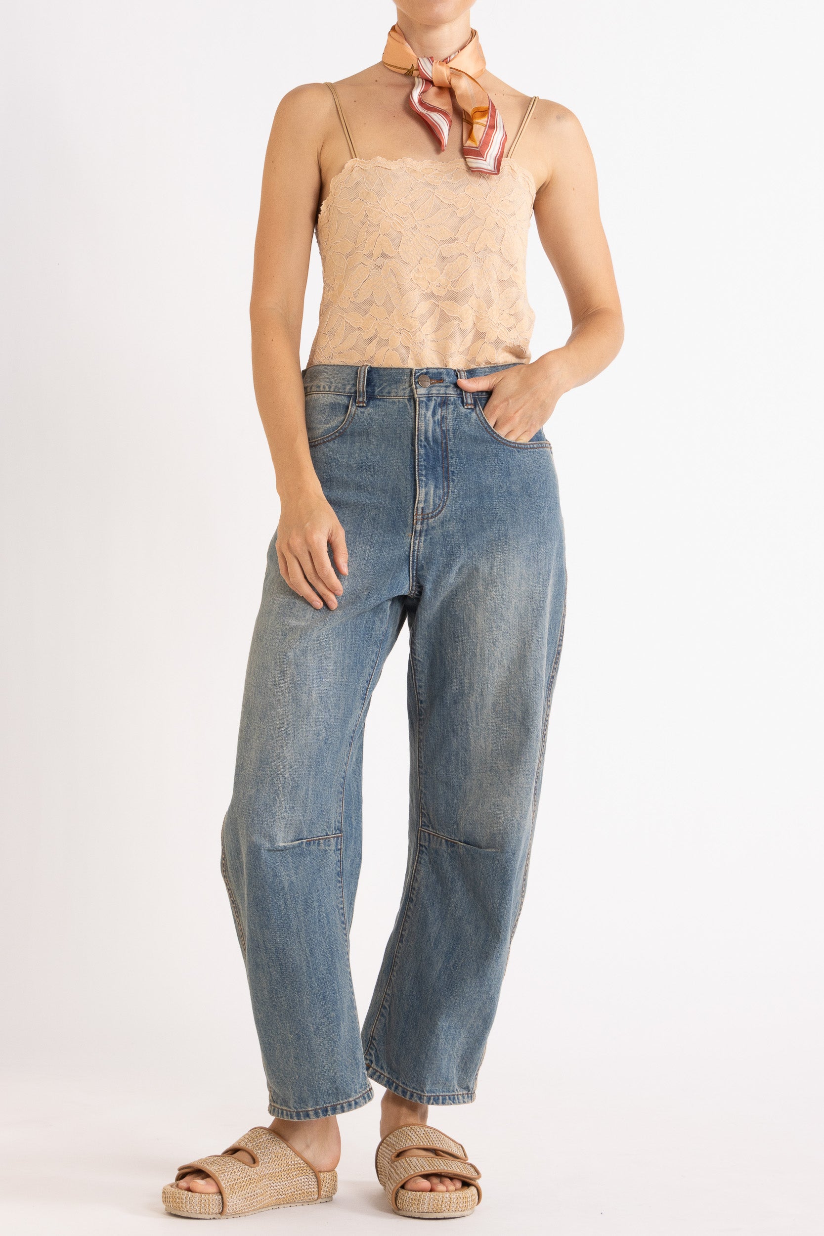 Person wearing a beige lace camisole, decorative scarf and blue jeans on a white background