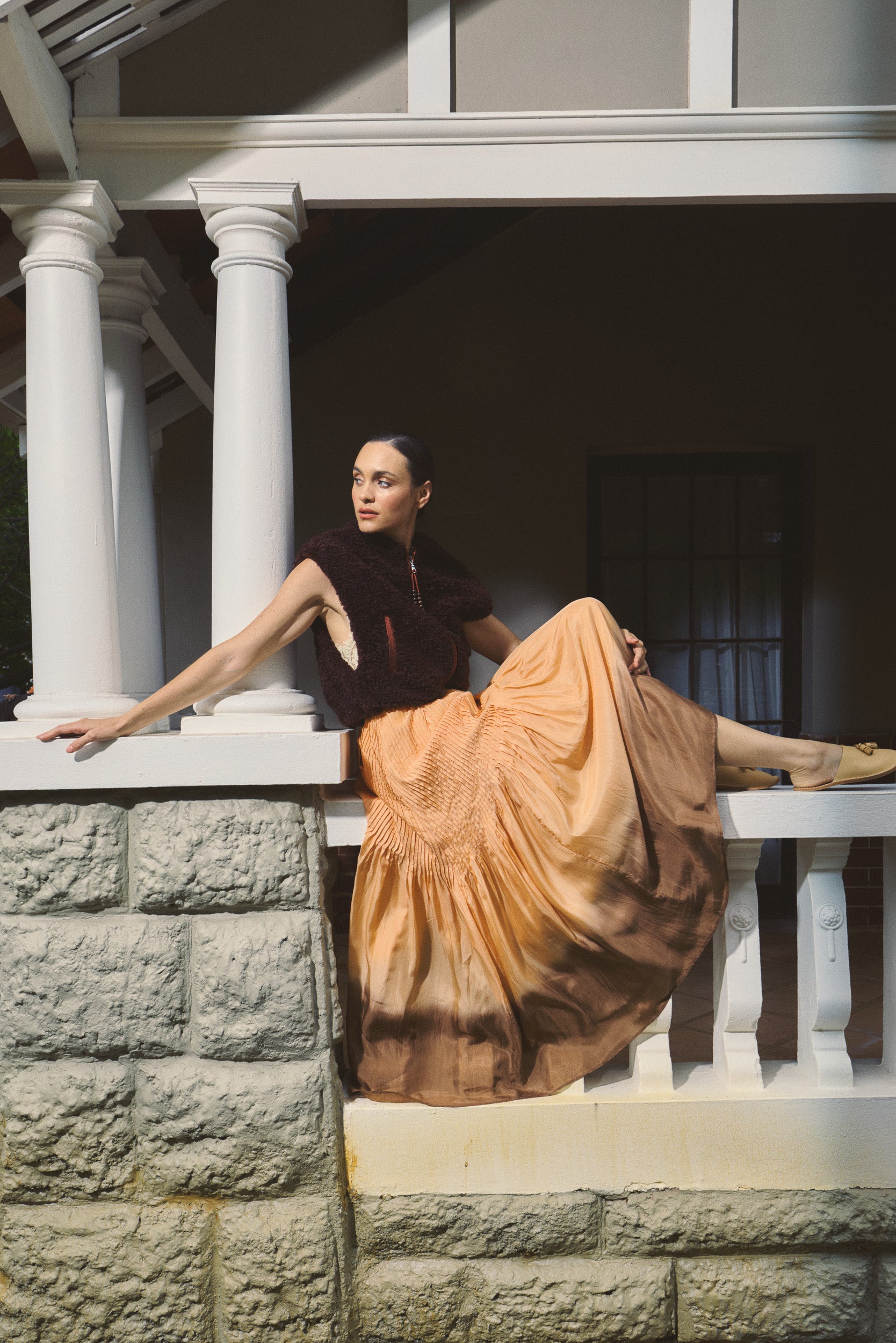Woman wearing port coloured faux fur vest, with a dip dyed silk habotai maxi dress in like orange, sitting on a balcony