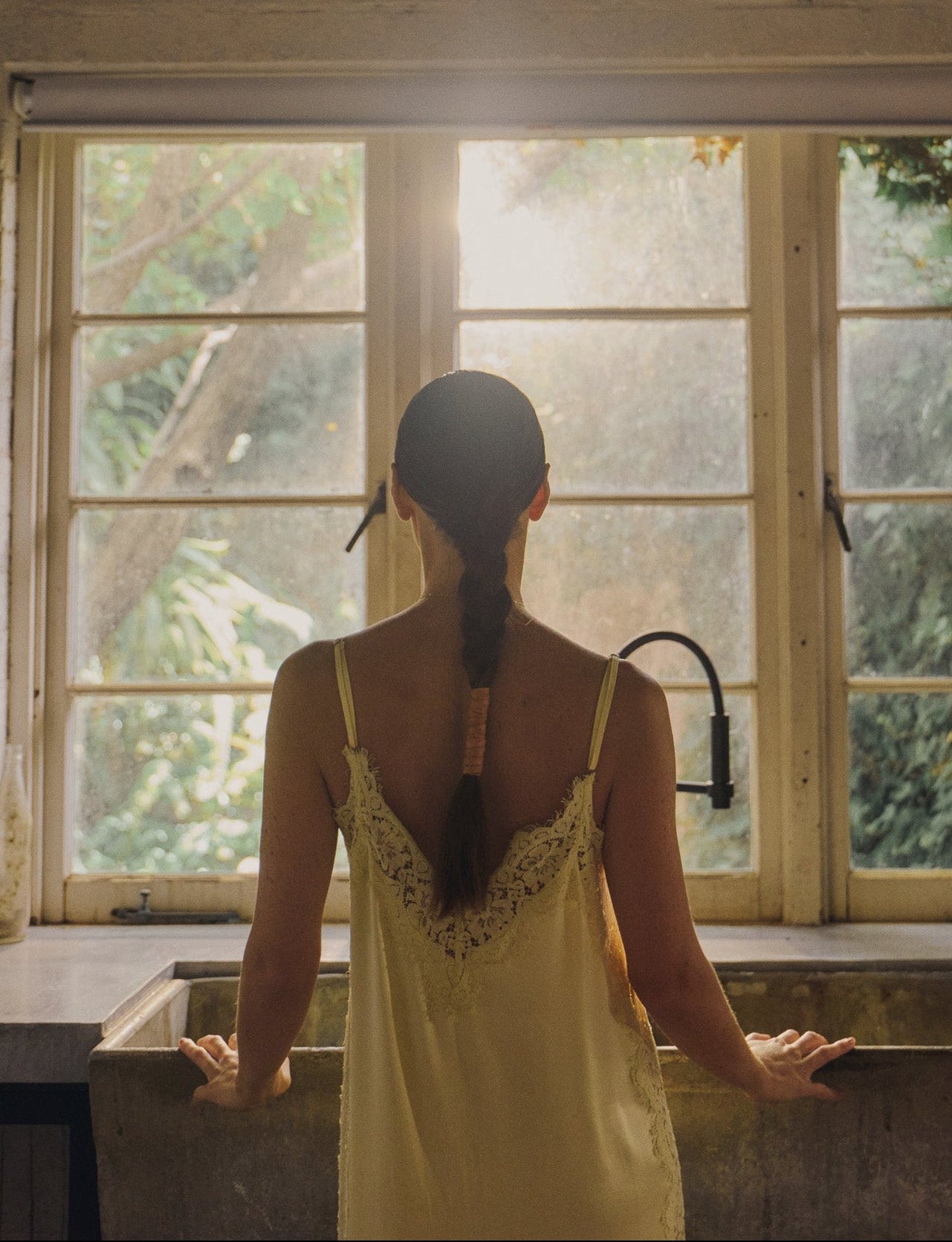Woman in a yellow dress standing in a kitchen with large windows, looking out at greenery.