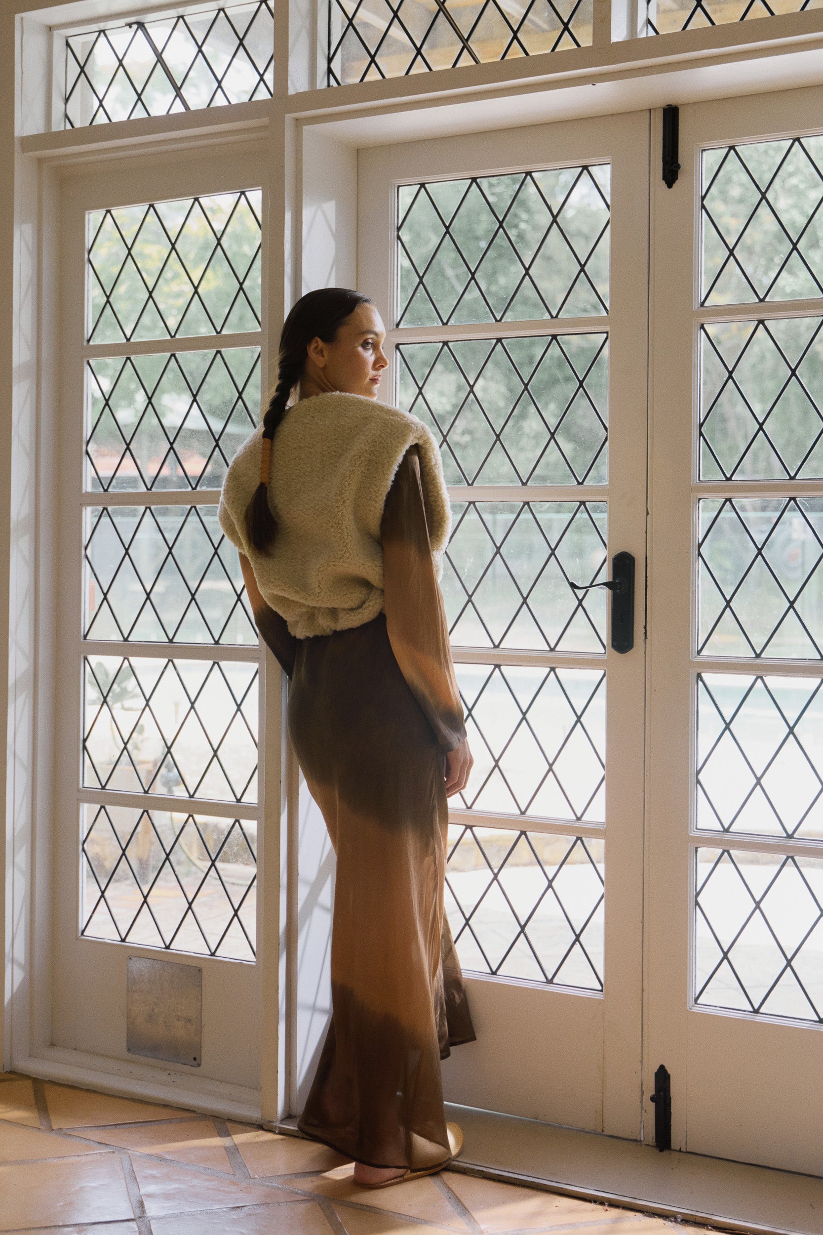 Woman wearing oat coloured faux fur vest over a long sleeve silk habotai maxi dress dip dyed in earthy tones, looking out of a window