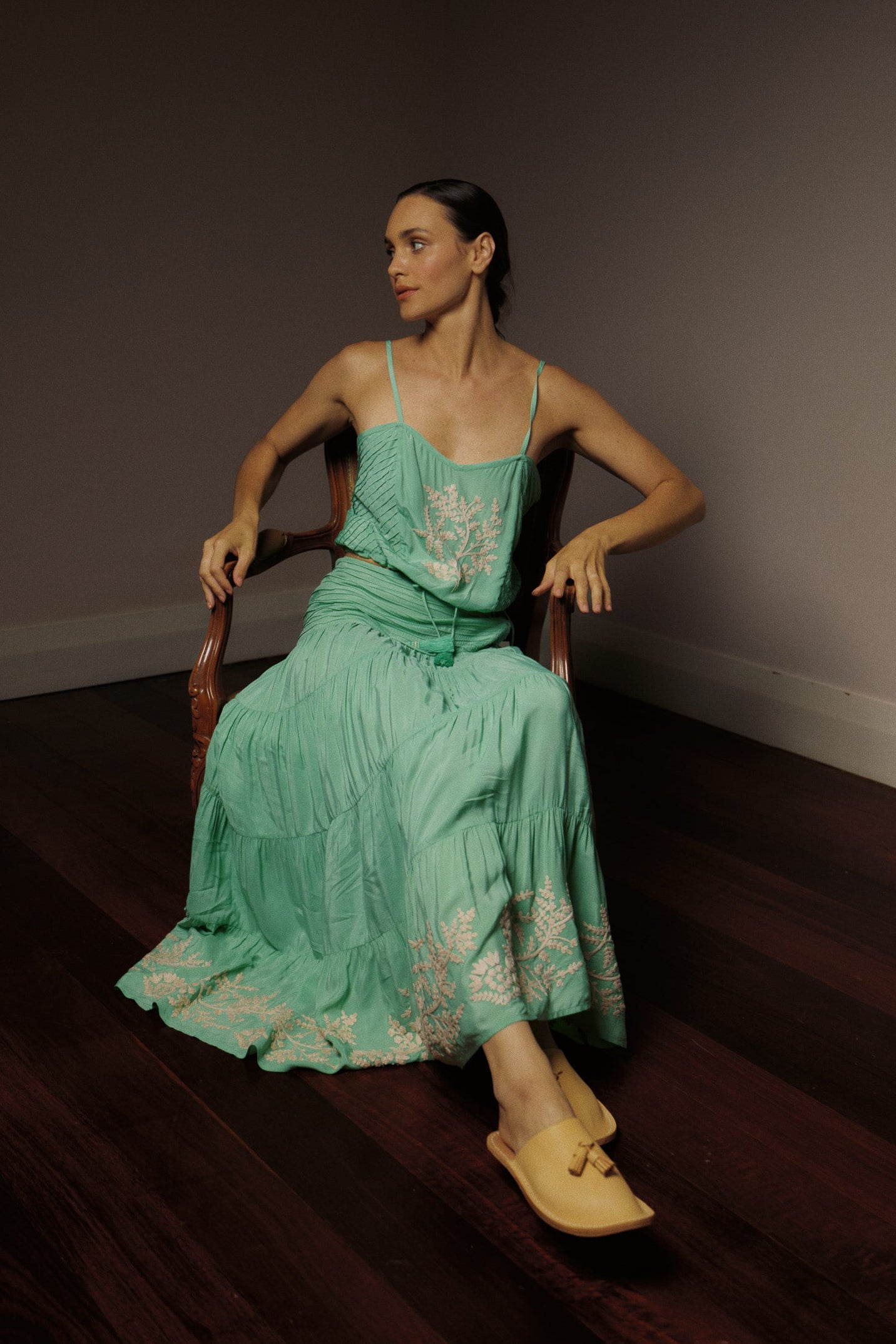 Woman sitting in a chair wearing a mint green camisole and long, tiered skirt with white floral embroidery