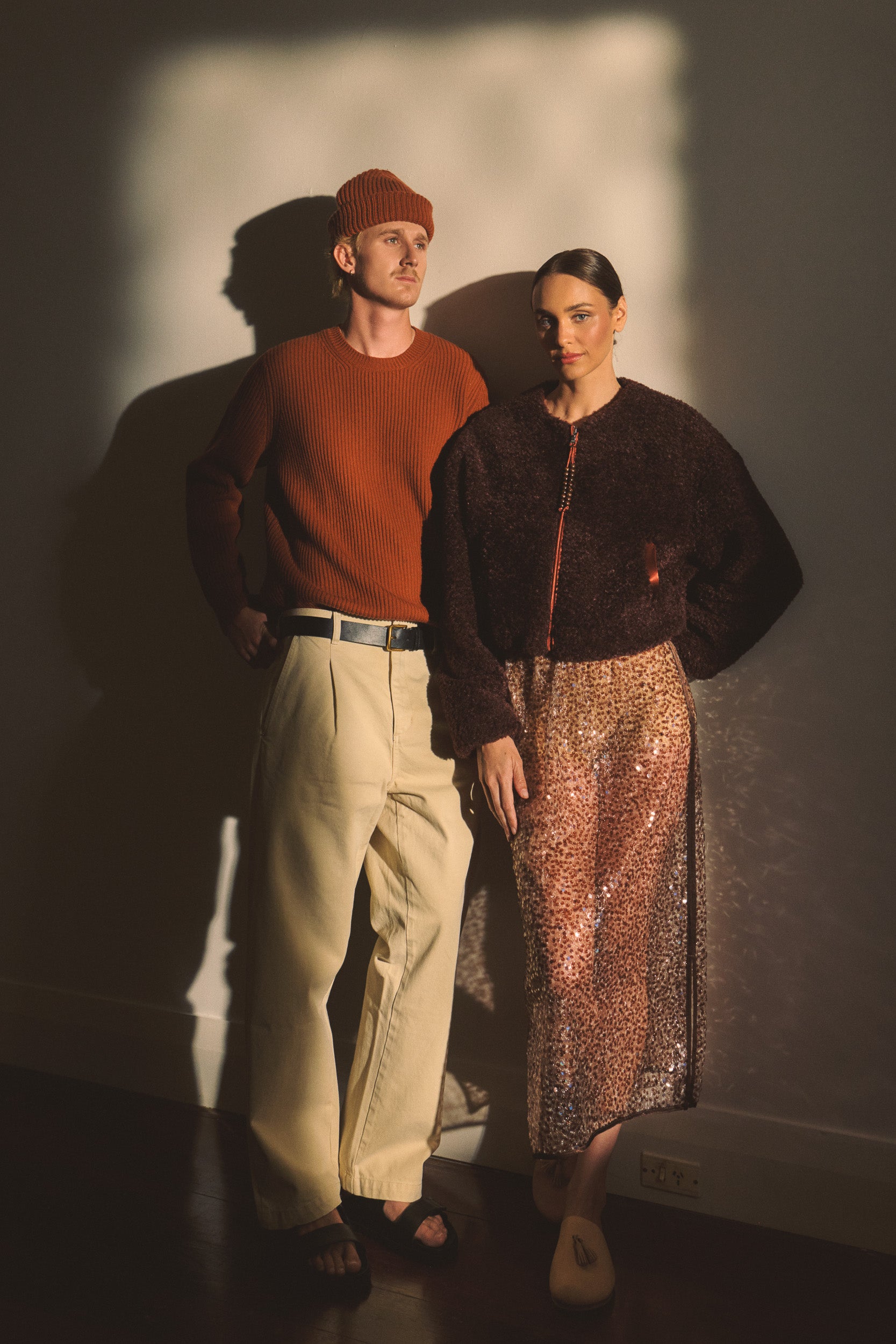 Male model wearing red clay beanie and sweater, with cream tailored pants. Female model wearing a port coloured faux fur jacket with a mocha sequin skirt. 