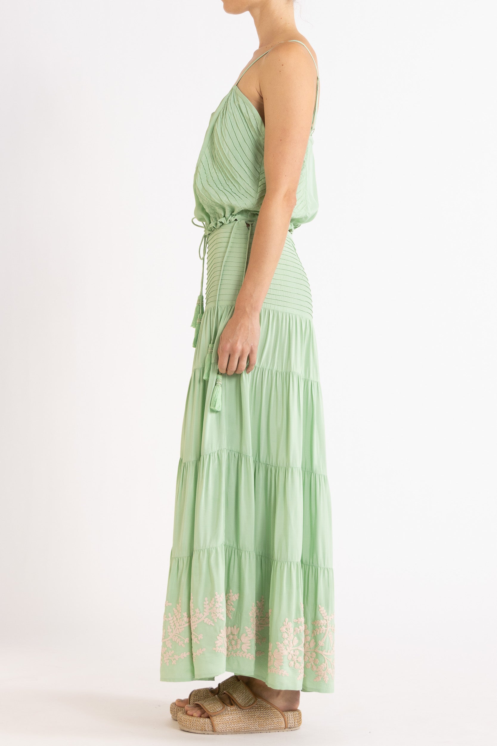 Woman wearing a mint green camisole and long, tiered skirt with white floral embroidery on a white background