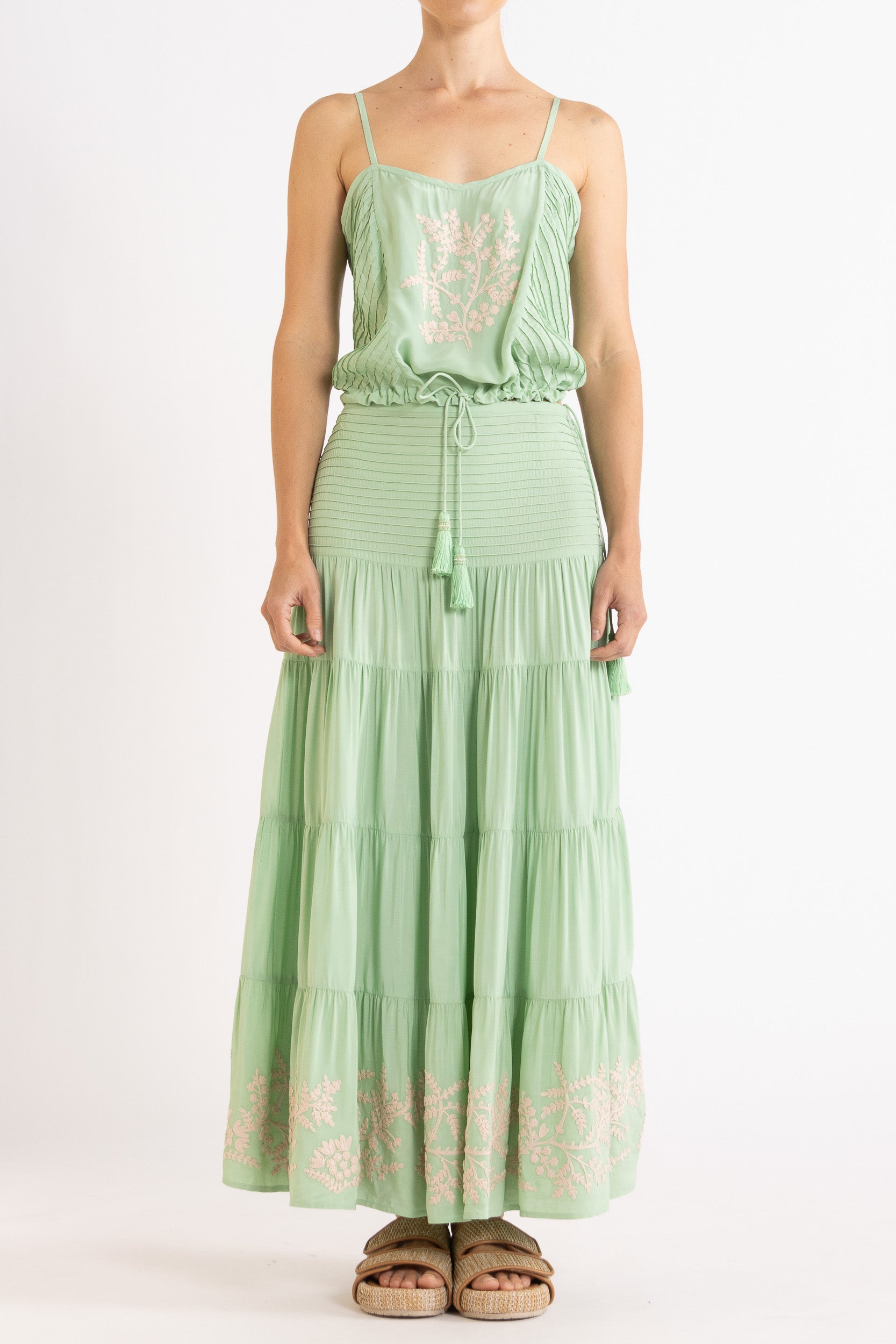 Woman wearing a mint green camisole and long, tiered skirt with white floral embroidery on a white background